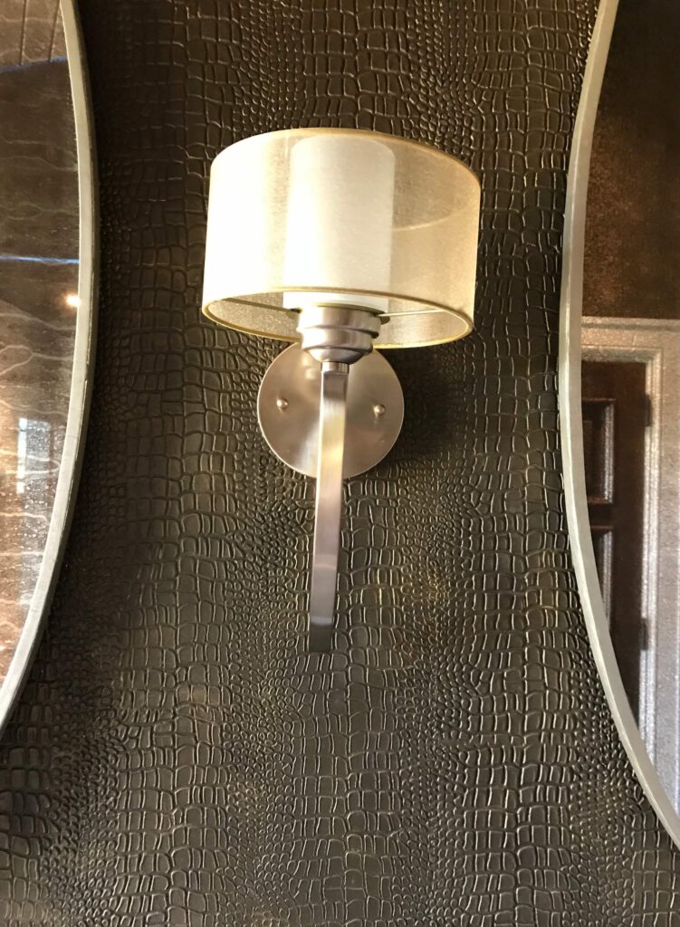 A close-up of a modern wall sconce mounted on a luxurious dark metallic gypsum plaster wall featuring a custom embossed crocodile skin texture in Westlake, Texas.