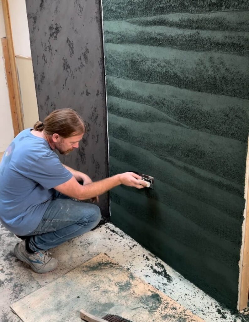 A photo of a male artisan plasterer in a workshop, hand-applying a steel trowel to a large green clay plaster wall featuring defined, undulating stratified textures. A second, darker mottled plaster panel is visible in the background, showing authentic craftsmanship.